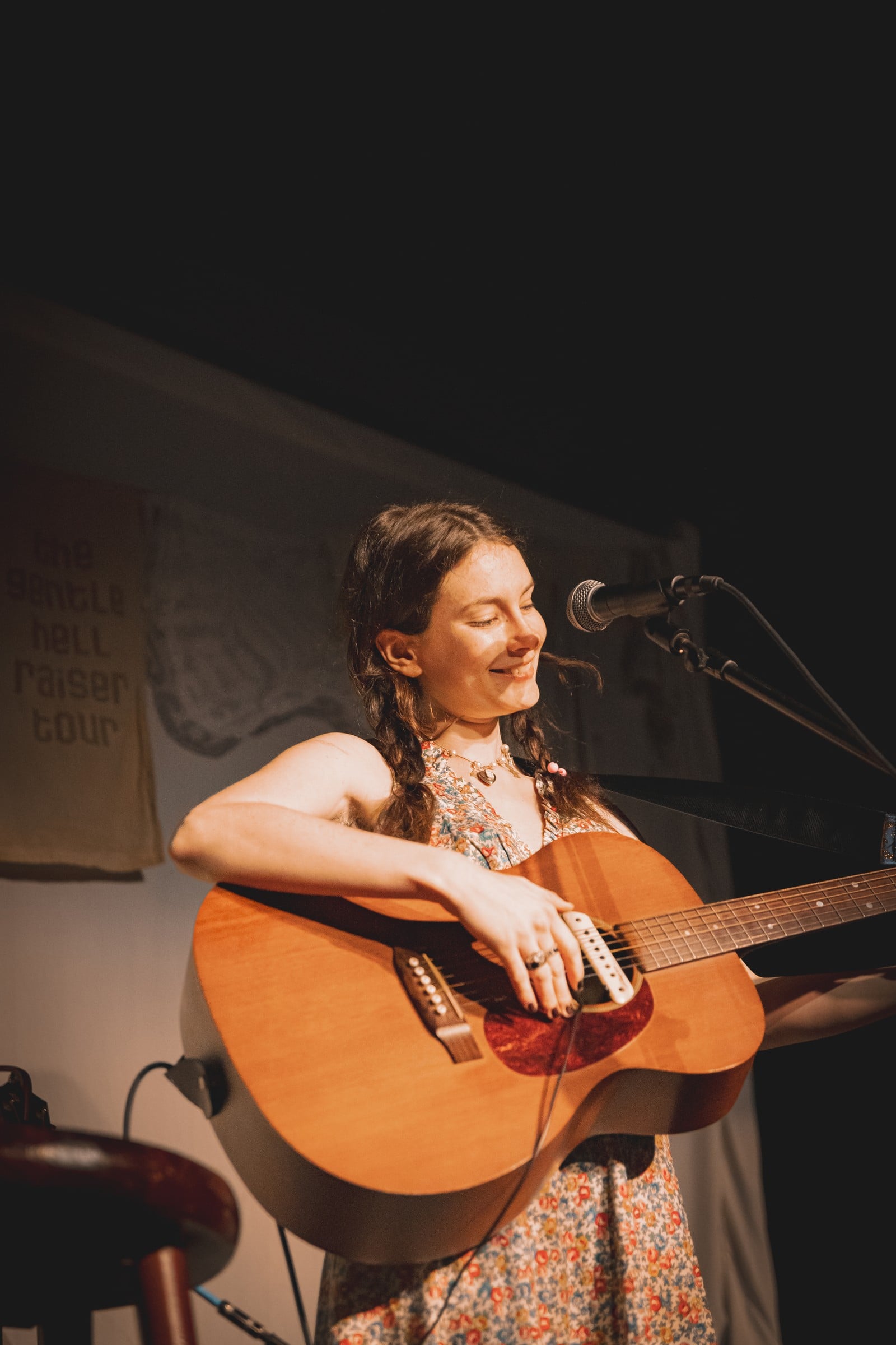 A performer holding an acoustic guitar beside a microphone under warm stage lighting.