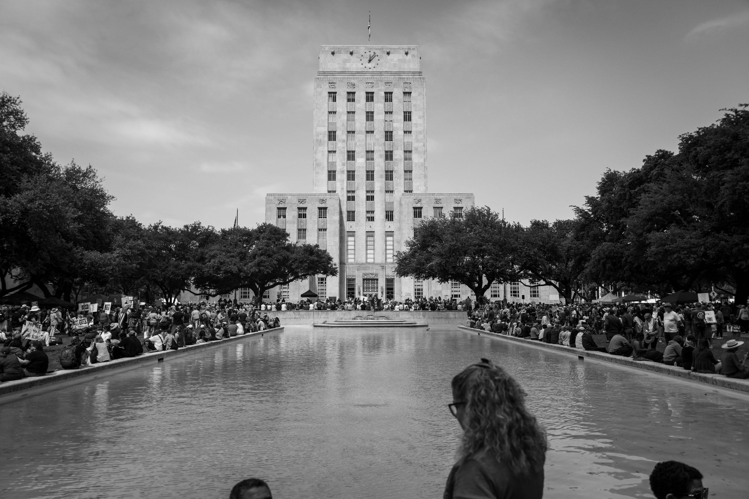 A crowd gathered around a reflecting pool in front of a tall civic building, in black and white.