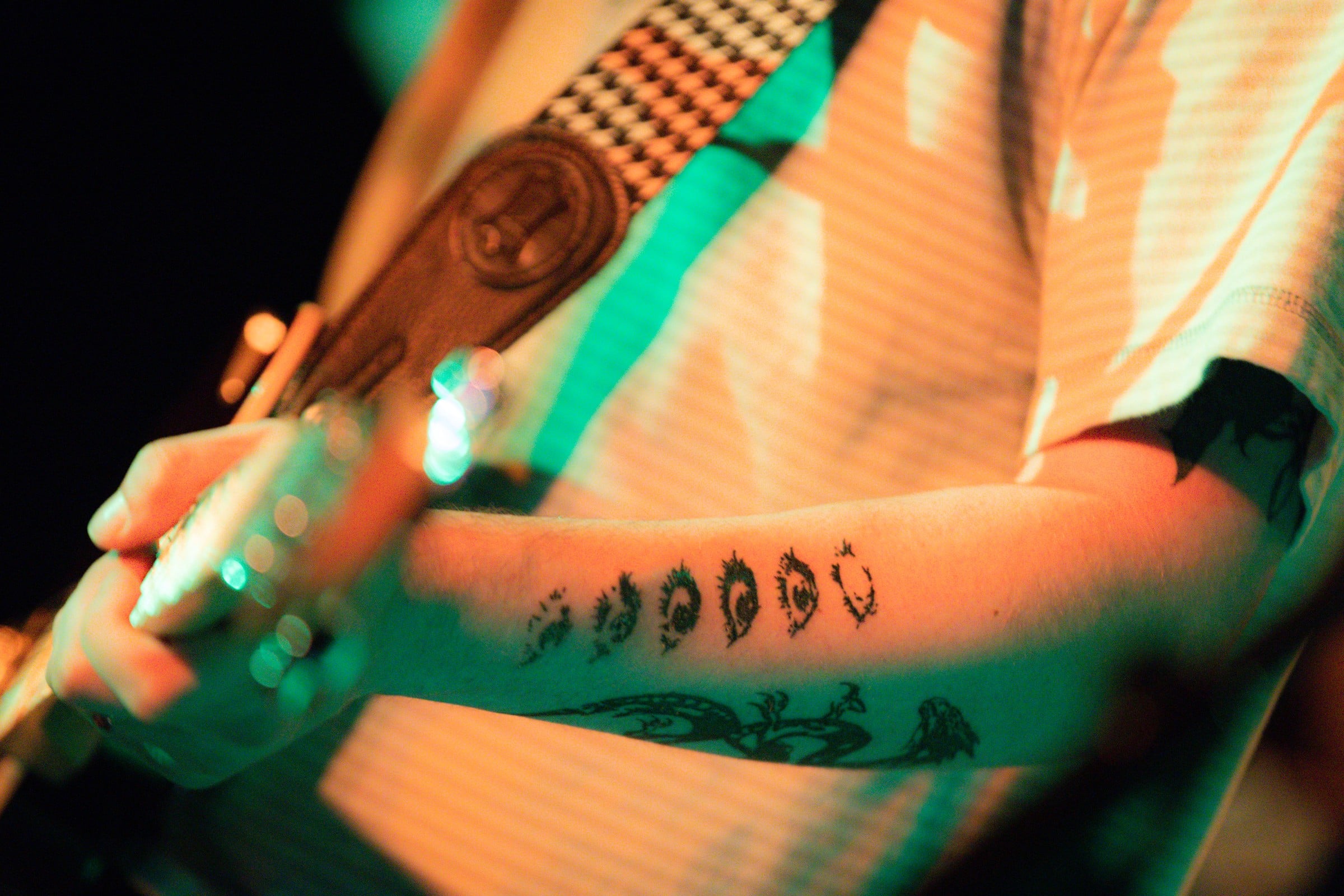 A close view of a tattooed arm and a guitar under warm and green stage lights.