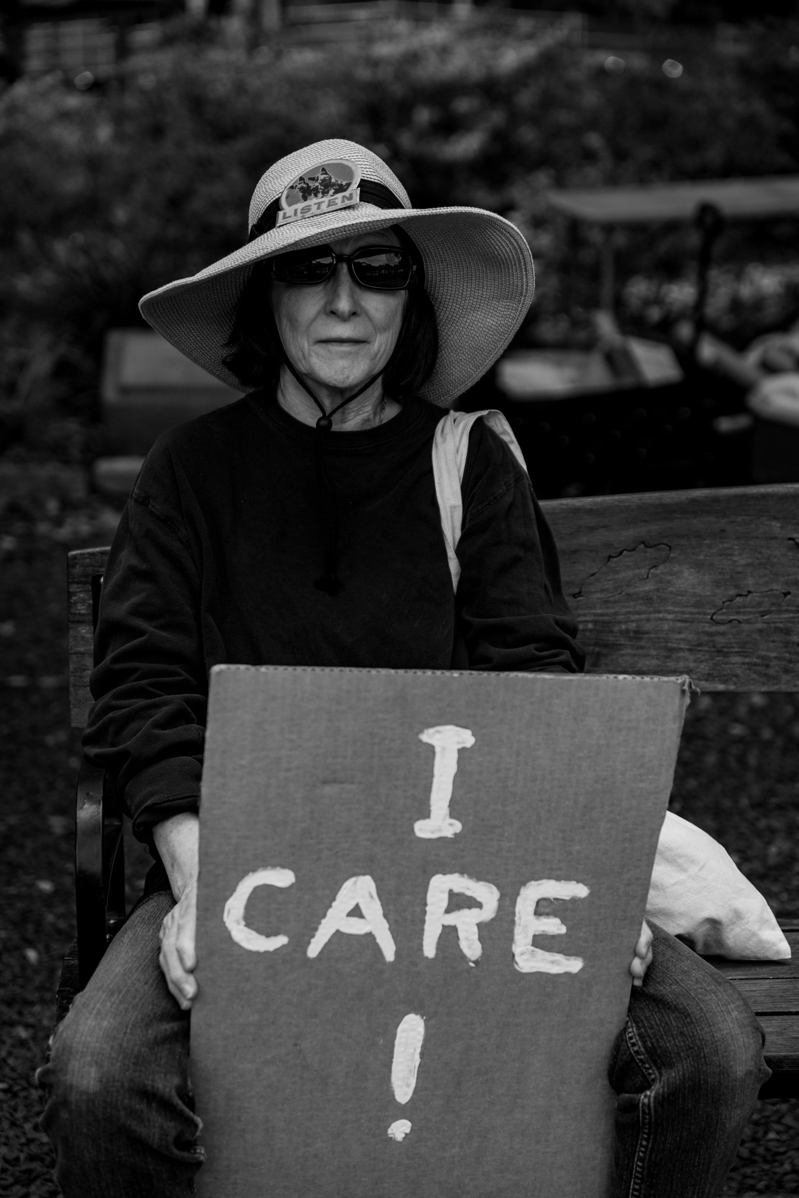 A black-and-white portrait of a seated person wearing a sun hat and holding a sign reading I CARE.