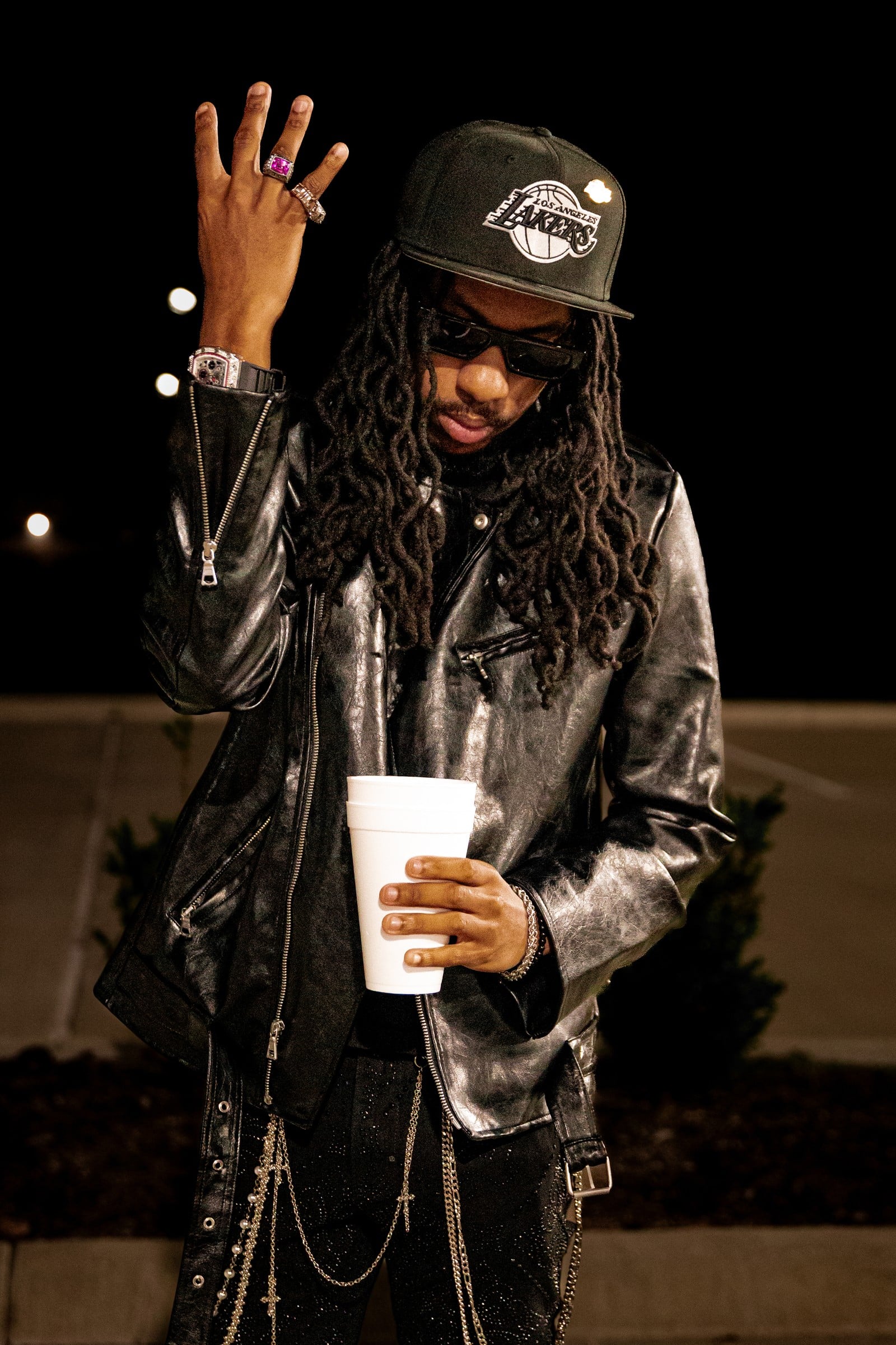 A nighttime portrait of a person in a leather jacket, cap and sunglasses, raising one hand.