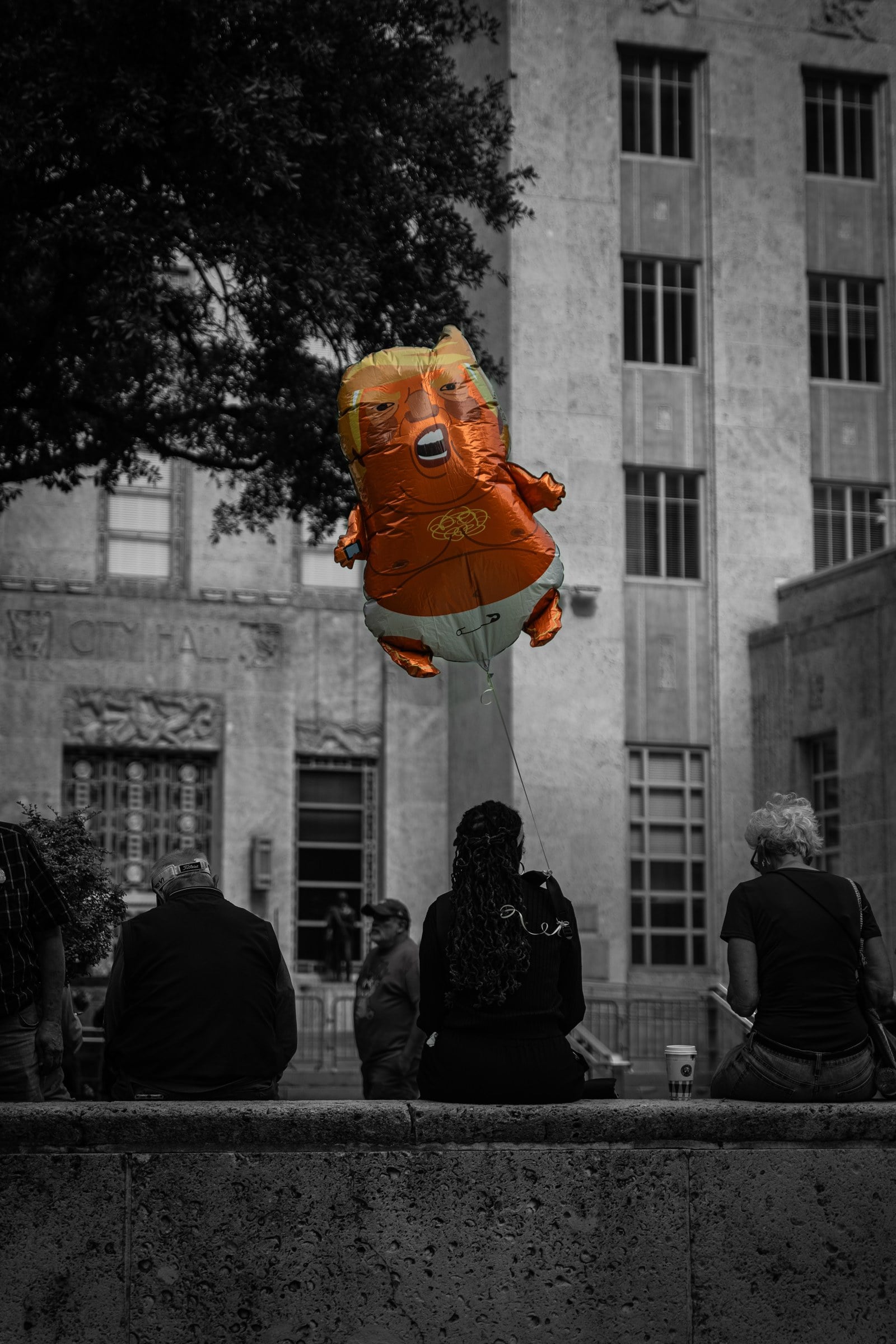 An orange character balloon above people sitting outdoors, with the surrounding scene in black and white.
