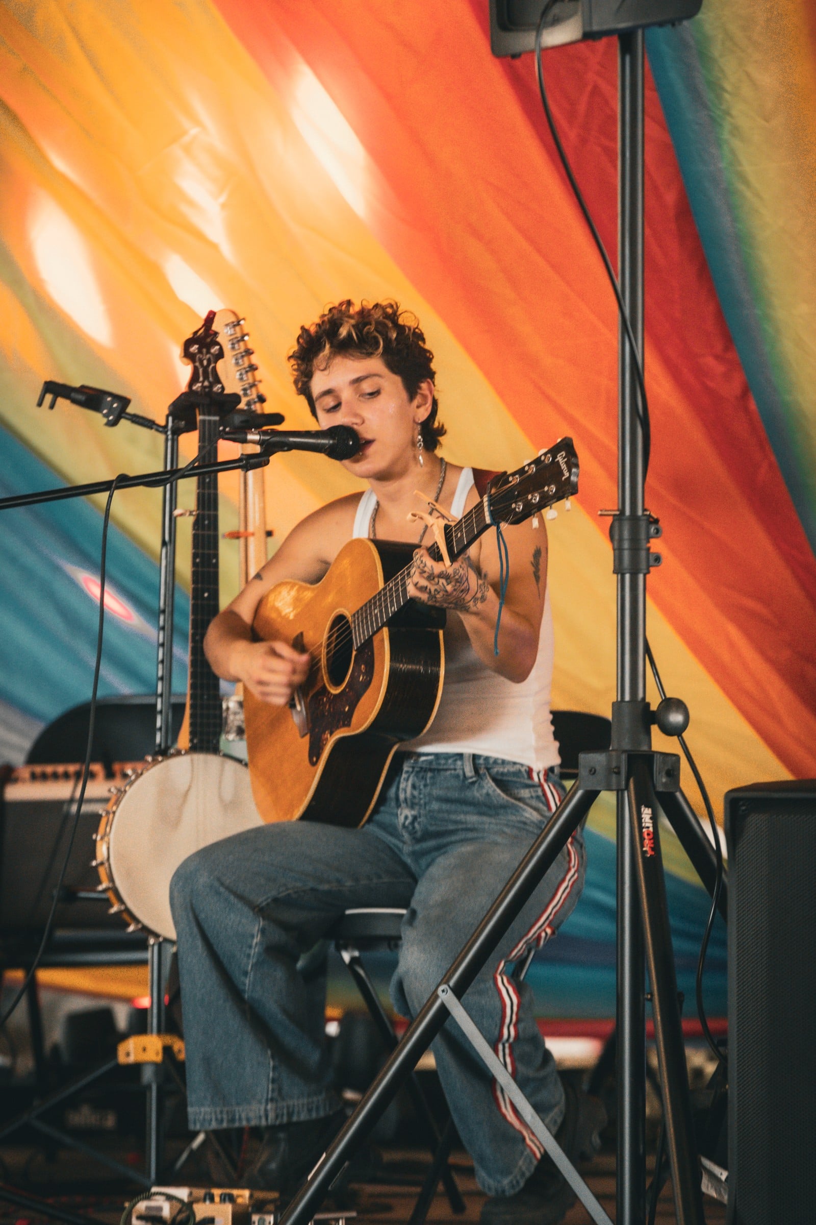 A seated performer singing and playing acoustic guitar against a colorful rainbow backdrop.