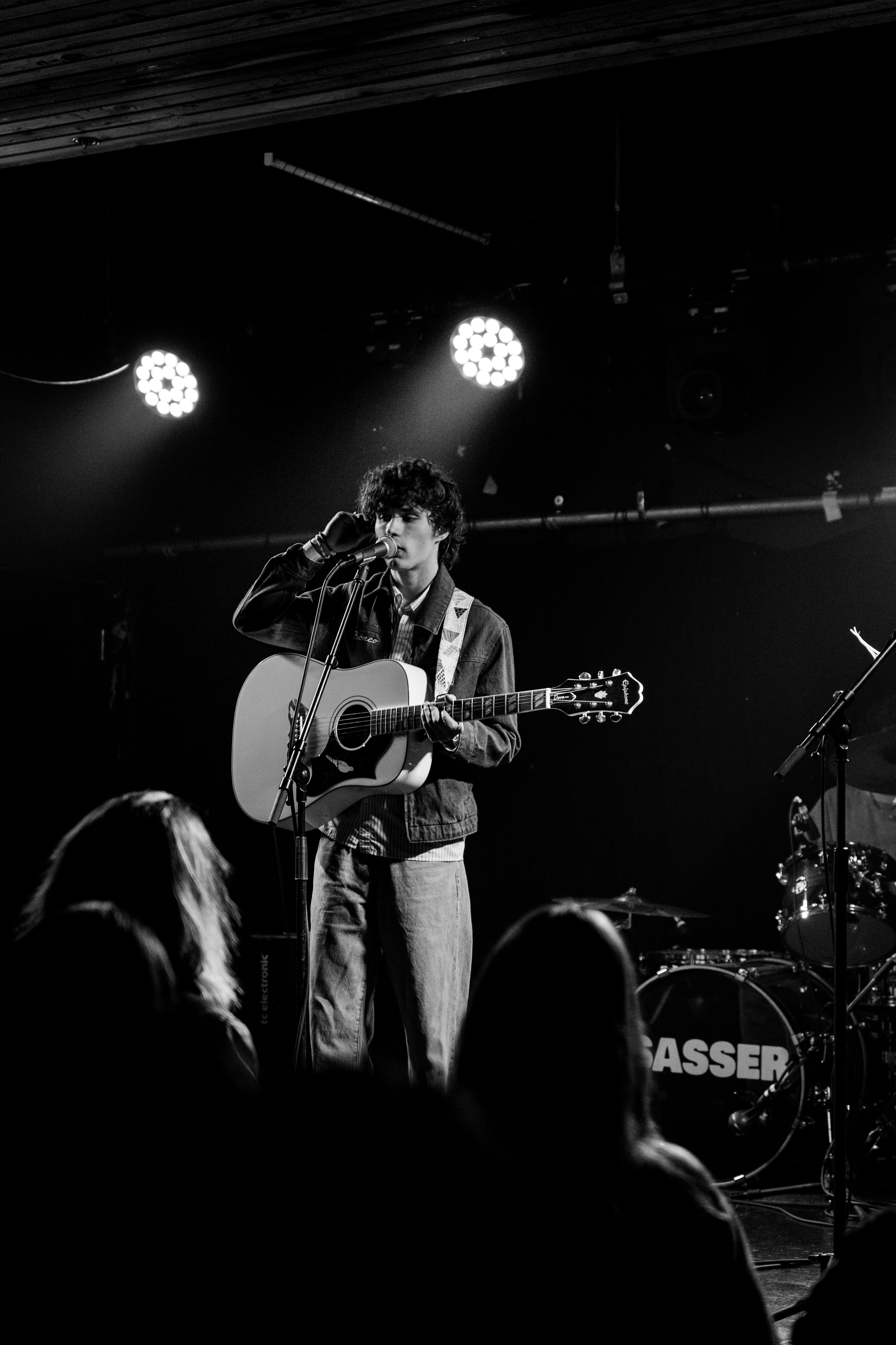 A musician singing into a microphone and playing guitar under stage lights, photographed in black and white.